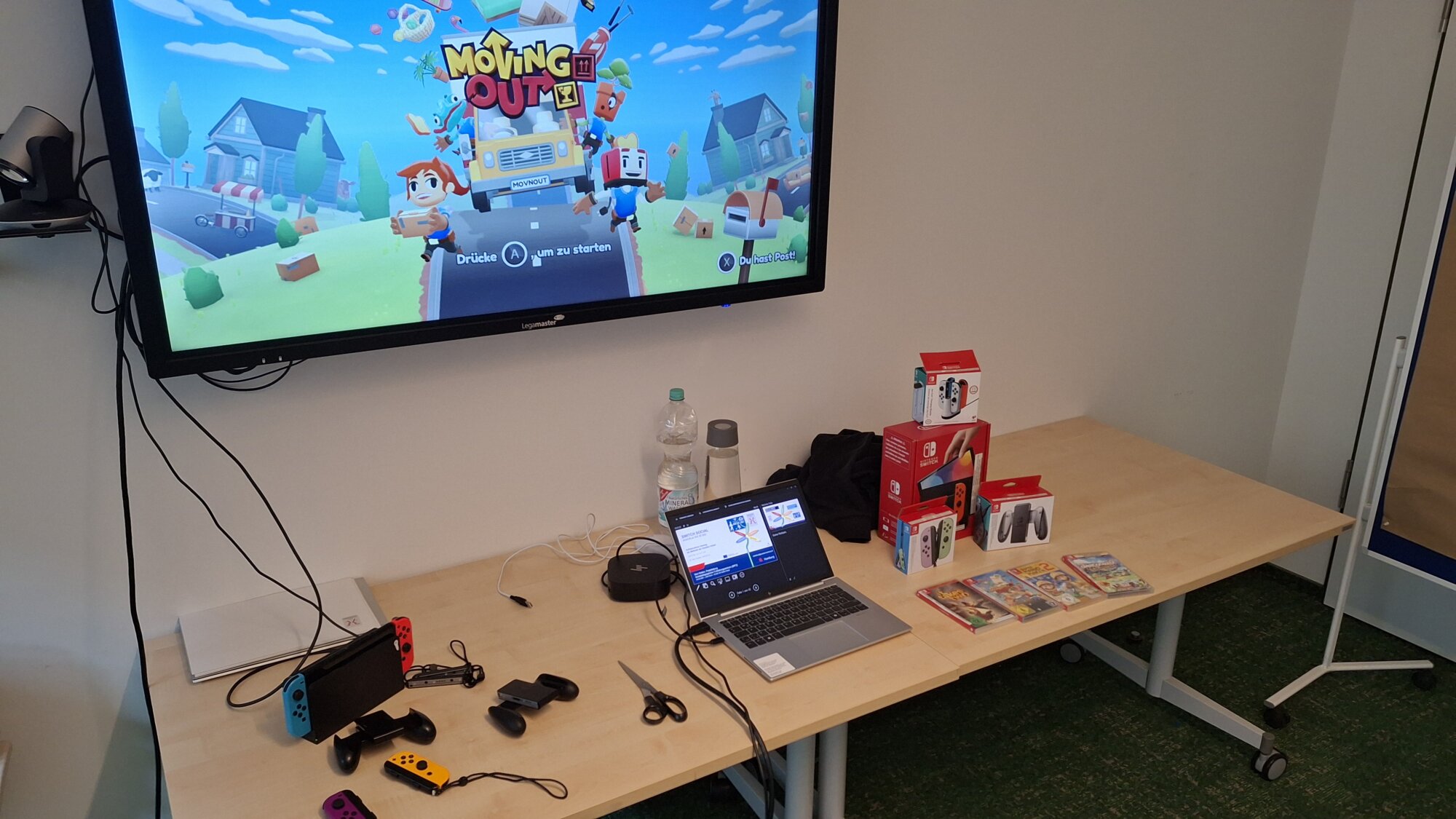 Setup für die Nintendo Switch im Kurs "Switch social". Auf dem Monitor im Hintergrund das Titelbild des SPiels Moving Out. Rechts vier Spiele und das Zubehör präsentiert für die Teilnehmenden. Links auf dem Tisch die Spielkonsole in der Dockingstation und zwei weitere Controller sowie Aufsätze.