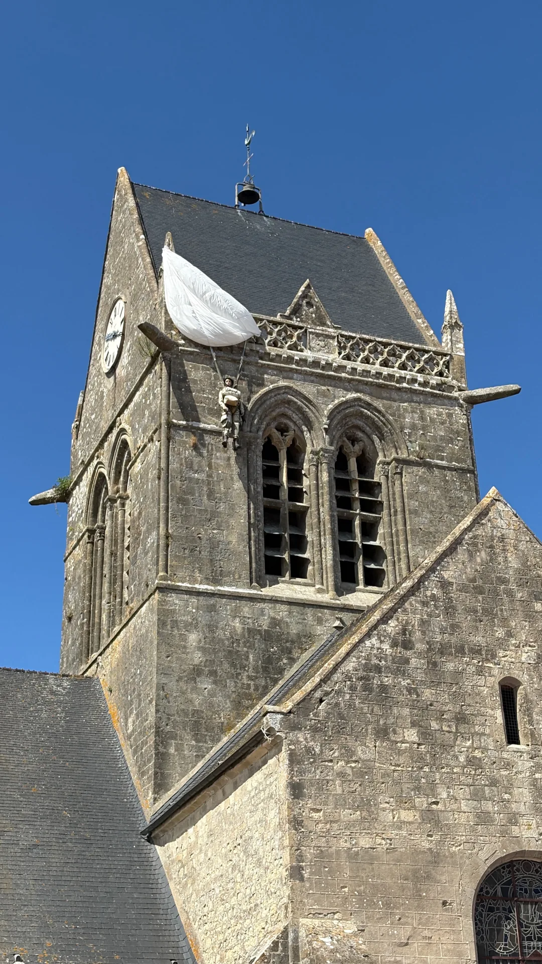 Vom Bildschirm in die Wirklichkeit: An der Kirche von Sainte-Mère-Église erinnert eine Fallschirmjägerfigur an John Steele, der in der Nacht zum 6. Juni 1944 mit seinem Fallschirm am Kirchturm hängen blieb. Foto: André Eymann, 2026.