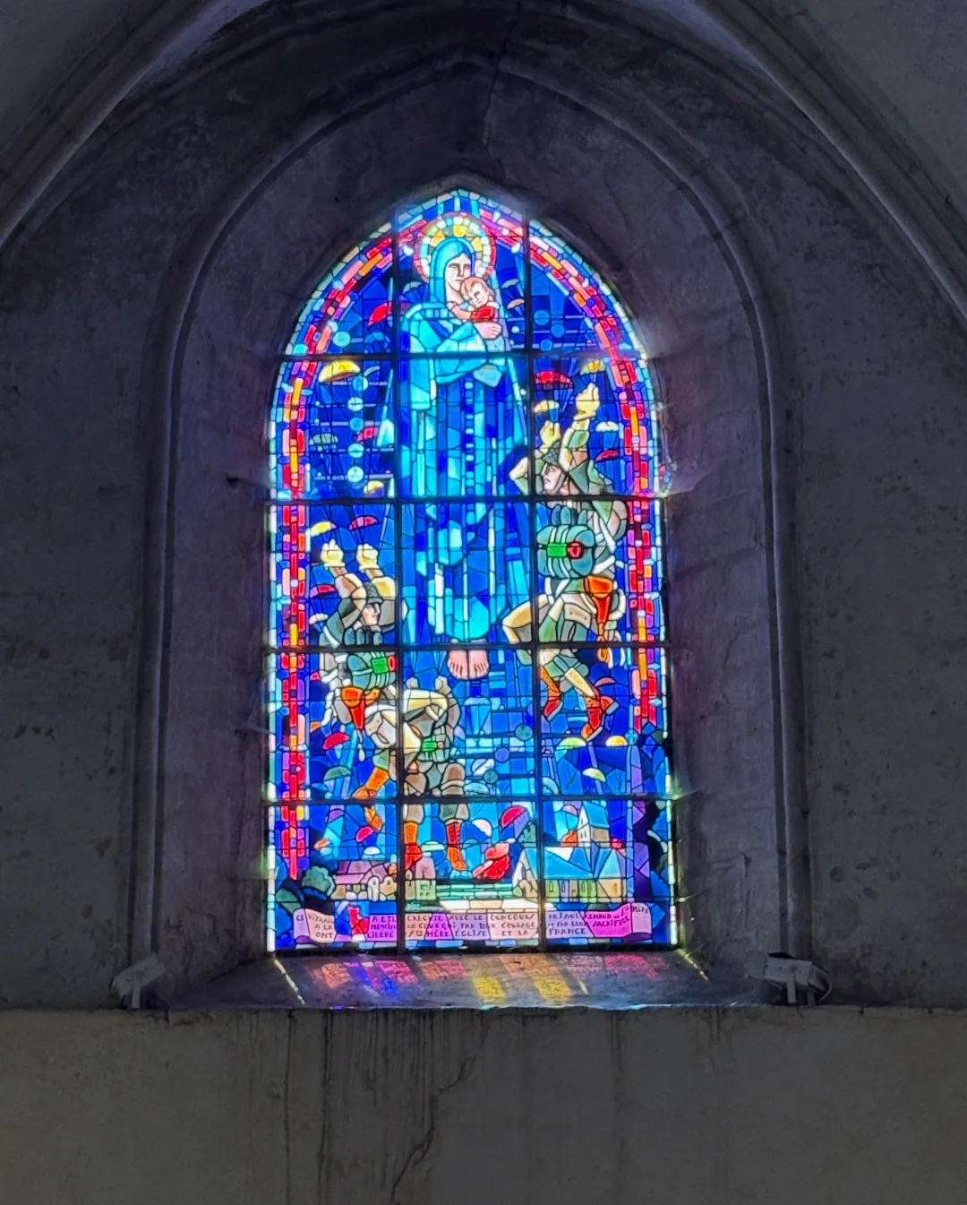Die Erinnerung reicht bis ins Innere der Kirche: Ein modernes Buntglasfenster zeigt Maria, umgeben von Fallschirmjägern, die über Sainte-Mère-Église herabschweben. Foto: André Eymann, 2026.